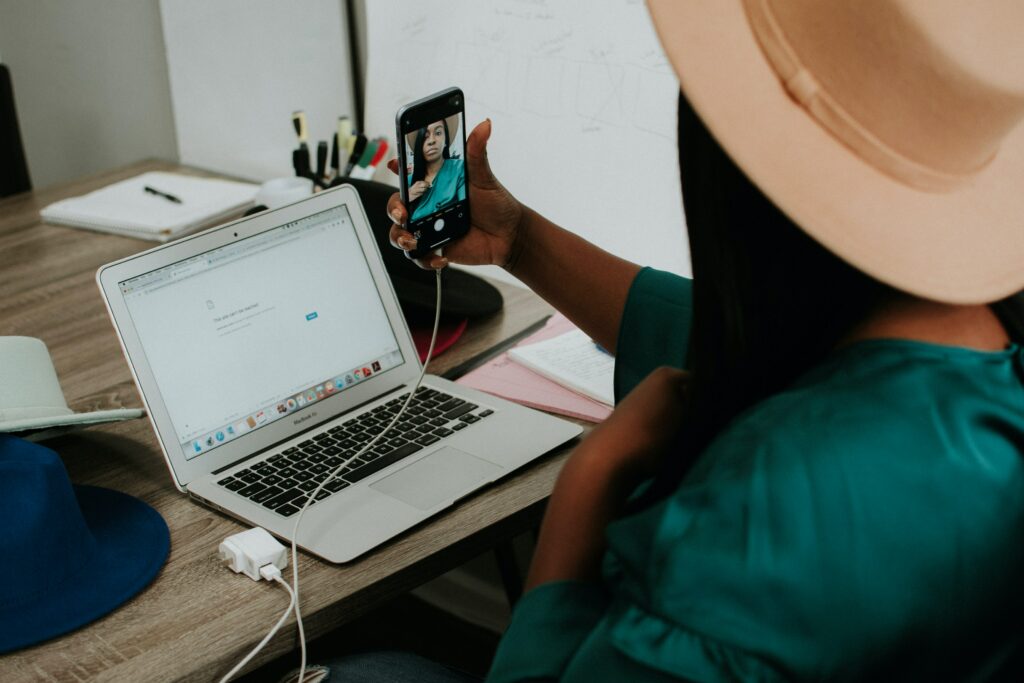 A freelancer holding up her phone and taking a selfie in front of her computer