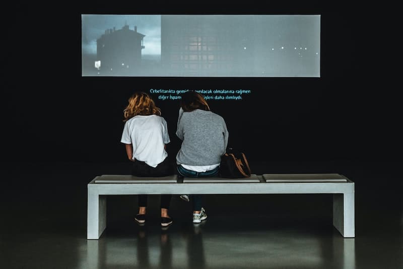 image of two women sitting in a museum while watching a video with subtitles