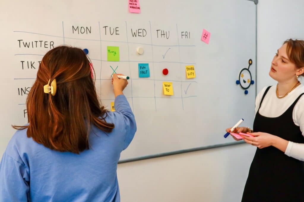 Two women working on a content calendar on a whiteboard with sticky notes