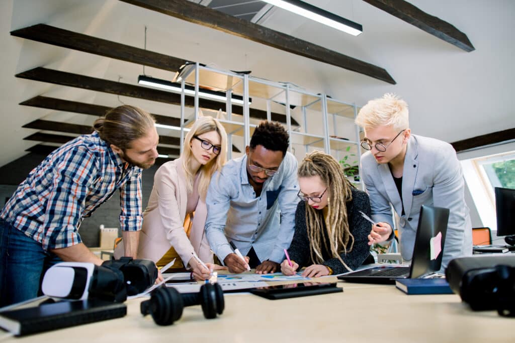 Creative multiethnic team of five skilled young colleagues working with data and marketing charts in modern office, leaning on the table.