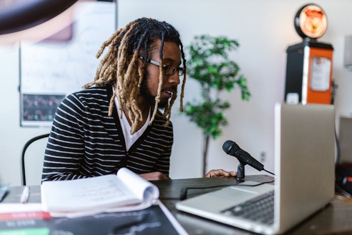 A young man with dreadlocks and eyeglasses records a podcast at a desk with a microphone and laptop.